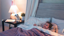 Happy young girl tucked into bed, smiling as she watches an educational projection on her ceiling from the Ozmotic Learning device.