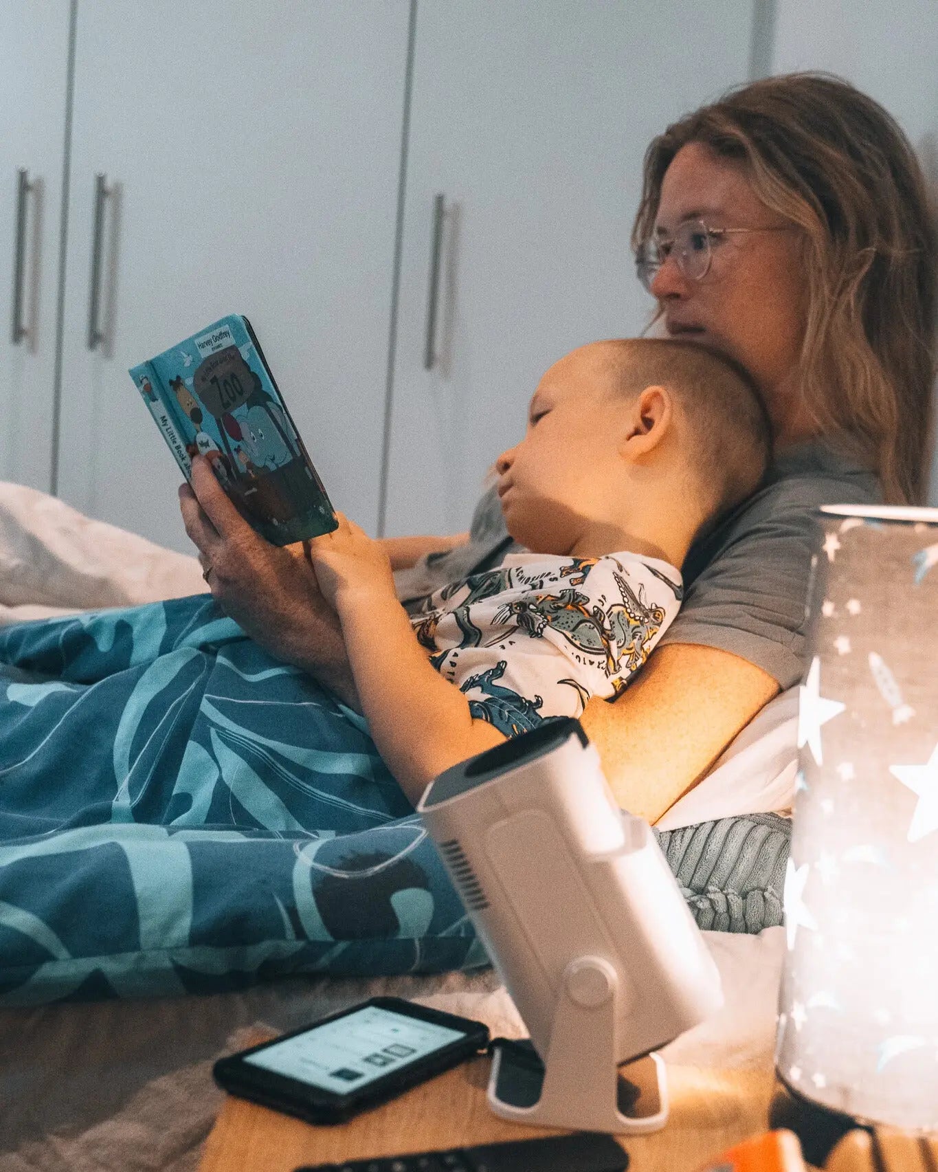 Mother reading a book to her child in bed, with the Ozmotic Learning projector resting on the mattress nearby.